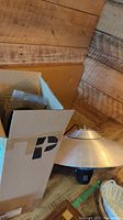 Photo of the large outdoor heater's conical aluminum reflector and nearby open cardboard box containing light fixture parts.