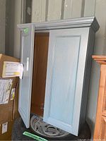 Photo of blue/grey wood medicine cabinet with door partially open, showing empty interior without shelves.