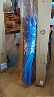 Full view of the wrapped umbrella and its long metal pole in packaging, resting against a large cardboard box