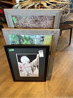Framed photos stacked, showing part of forest and porch scene photos, and a black framed poster.