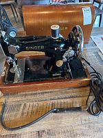 Full view of the vintage black Singer sewing machine with knee pedal and wooden case cover marked 'Singer'. The machine rests on a wooden base with all parts visible including power cord and pedal.