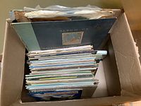 View looking down into a cardboard box packed with approximately 40 vinyl albums mostly stacked vertically showing various album cover edges and jackets.