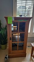 Front view of the oak cabinet showing the glass front door sectioned into four panels, black latch handle, and some visible scratches on the wood surface.