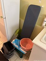 Ironing board leaning against wall next to refrigerator and sink with ironing sleeve board, green ironing pad, black woven baskets, plastic tub, hand broom and dustpan on floor.
