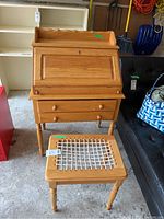 Front view of wooden secretary desk with fold-down desktop and front drawers, paired with wooden stool with white webbed seat.