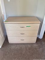 Front facing photo of light wood dresser with three drawers and silver handles on carpeted floor.