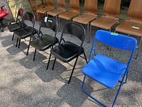 Four black metal folding chairs and one blue IKEA folding chair positioned outdoors on pavement. Chairs show minor scratches and wear.