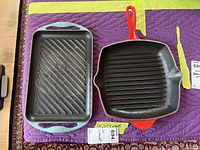 Two Le Creuset cast iron grill pans on purple quilted surface: one red square pan with ridges and one light blue rectangular pan with ridges.