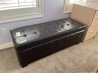 Perspective view of dark brown leather rectangular bench ottoman with tufted top, positioned in front of window, placed on carpet.
