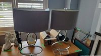 Pair of two brushed metal and acrylic table lamps with oval gray fabric shades displayed on a wooden table near window light.