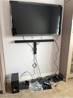 TV mounted on wall with soundbar below and subwoofer and Blu-ray player on floor with cables and remotes.