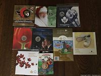 Photo of multiple Royal Canadian Mint coin sets and collections in original packaging laid on a wooden surface, including 2006 Baby Steps, 2006 Canada train themed, Maple Leaves 2010, 2007 Classic Classiques, and sports-themed items.