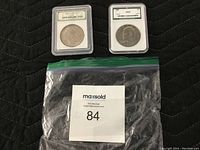 Both graded silver dollar coins displayed in their protective plastic holders with grading certificates visible, bags and lot tag in view.