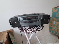 Black Memorex dual cassette player/radio with dual cassette decks, radio tuner, and speakers on both sides, placed on a small stool with a patterned cover and boxes in background.
