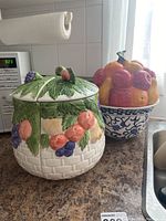Photo of two ceramic jars on a kitchen counter near a microwave and sink. The foreground jar is shorter with fruit and leaf designs and a segmented lid. The background jar is taller with a fruit-shaped lid and blue and white base.