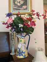 Ceramic pitcher and two tall mirrored mosaic vases with faux flowers arranged together on a wooden surface under a framed floral painting