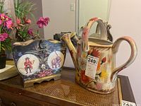 Two-piece lot with ceramic watering can and decorative box on a table with floral arrangement in background