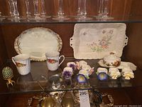 A shelf with two serving trays, two mugs, a decorative egg, floral napkin rings, covered butter dish with floral pattern