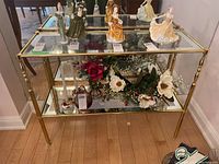 Console table with brass frame and glass top and bottom shelf. Displayed with decorative figurines and artificial flowers (not included).