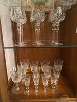 Wide shot showing two shelves of various styles and sizes of crystal stemware and glasses, all clear crystal with cut patterns, some glasses showing wear or chips.