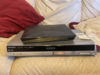 Panasonic DMR-ES20 DVD recorder with front panel and Sony Blu-ray player stacked on top, placed on a beige leather couch with pillows in the background.