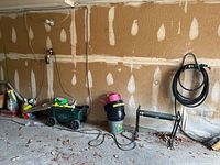 Garage wall storage of garden supplies showing Lawn Buddy garden cart, Yardworks folding stool/kneeler, coiled garden hose on hook, and black metal yard edging piece. Floor dusty with debris.