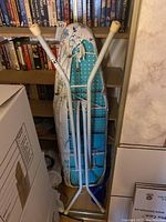 Photo showing folding ironing board with blue and white dotted surface leaning against a bookshelf, with smaller sleeve board attached to it