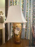 Front view of the tabletop lamp showing floral and bird motifs on the porcelain base and the white square lampshade with tassels.