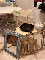 Photo showing Mid-Century Modern chair with cream-colored curved backrest and seat, cast iron frying pan on the chair seat, rectangular wall mirror and several picture frames on the floor in front of the chair.