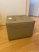 Photo showing front and side angle of a small vinyl square ottoman with tufted top and piping along edges on wooden floor.