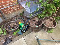 Three matching large outdoor planters with dried plant material inside, placed on a patio area near a brick wall and ivy. Accompanied by gardening tools including a weed puller and sprinkler.