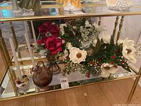 Wide view showing the glass bowl with faux flowers, the small brown vase, and floral faux arrangements on a glass shelf.