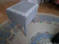 Floral patterned sewing seat with blue tapered legs resting on a floral rug. Seat is square with lace trim on top edges.