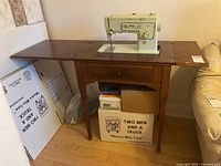 Singer Stylist sewing machine mounted on wooden table with fold-out extensions, showing general view and condition.