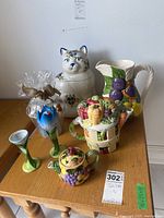 Full view of the lot on a wooden table showing ceramic cookie jar in the shape of a dog's head, ceramic pitcher with fruit decoration, teapot, candle holder, and wrapped glass vase.