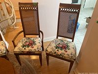 Two wooden cane-backed dining chairs, one captain-style with armrests and one side chair without arms, showing floral cushions and circular cutout pattern on chair backs.