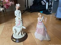 Pair of Wedgwood ceramic figurines: a dog walker with white dog on wooden base and a woman in an elegant pink and white gown with fan, positioned on a hardwood floor.