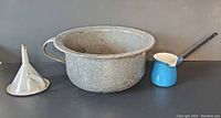 Three enamel kitchen items: white funnel with blue rim, white chamber pot, and blue butter warmer with ladle placed on a dark surface.
