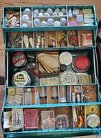 Open tackle box showing multiple compartments filled with assorted vintage fishing lures, packaged hooks, split shots and spools of fishing line