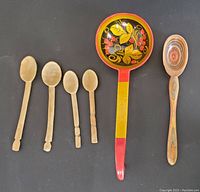 Photo of all spoons together on black background showing 4 antique primitive hand carved spoons, a Norpro rainbow wooden spoon and a vintage Russian hand painted ladle.