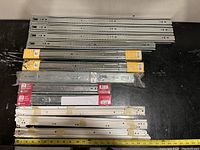 Overhead photo of various metal drawer slides on black surface with measuring tape showing length, some still in original packaging.