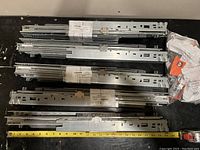 Five metallic silver steel drawer slides, each approximately 600mm long, laid out on a black surface with measuring tape indicating length. Some slides have original labels.