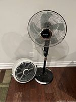 Photo shows two fans - one tall black standing fan with a metal blade design and one shorter white floor fan with four blades.