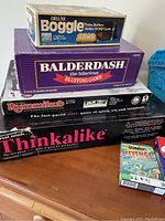 Photo showing stacked game boxes including Deluxe Boggle, Balderdash, Rummikub, Thinkalikе, and a small card game on a wooden surface