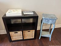 Front view of black IKEA 4 compartment storage unit with 2 BRANAS wicker baskets and small chalk painted side table with drawer missing knob