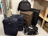 Photo showing six luggage pieces: two navy blue wheeled suitcases, one brown wheeled suitcase, one black wheeled suitcase, a black carry-on bag, and a black duffel bag arranged on floor next to shelving.