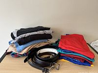 Two stacks of men's clothes, one of sweaters in neutral and darker colors, another of sport/golf shirts in red, blue, teal, yellow, maroon, with several leather belts coiled beside.