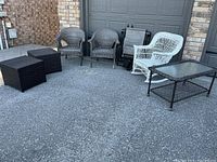 Photo shows the full set of outdoor furniture arranged on driveway: a white wicker rocker, two grey wicker armchairs, two black cube side tables, one rectangular coffee table with glass top, and two folded black loungers stacked.