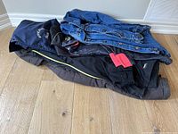 Stack of various jackets including windbreakers, winter coat, and denim jackets on wood floor.