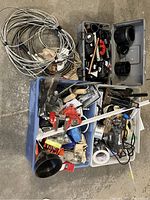 Photo showing bins filled with various plumbing parts including multiple black plastic pipe fittings and metal couplings, coiled metal cables or wires, and assorted small hand tools.